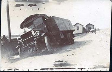 The image shows a Chevrolet truck on the side of the road leaning badly to the left.  A man can be seen to the left of the photograph near a pole.  There is a fishing shed plus another shed in the background.  Some men and a cart can be seen near the fishing shed.  