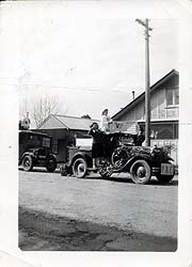 This image shows two cars driving down Salmon Street. The first car is festooned with branches and has one young woman standing on the roof holding a sign reading “V.P” A man is standing on the left running board also holding a sign reading “V.P” and another young woman is standing on the right running board. The car behind shows only the front part and has people standing on the roof holding a sign depicting a cross. A  single story timber building is in the background and next to it behind the car is a double story timber building on which the word “Corner” can be seen. A wooden power pole is in front of this building.