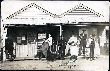 The image shows a double fronted single storied timber building with a corrugated iron roofed verandah. Above a window in the building is a sign on which the words “Hairdresser Tobacconist” can be made out. There is another sign which reads “The Peninsula Post ... sold here”. Leaning against the verandah is a bicycle. In front of the building are, from left to right, a man leaning against a verandah post, a man seated with another man standing behind him cutting his hair, a man seated with a football, a man standing holding a cricket bat, a man standing in a white coat holding a doctor’s bag, a man standing holding an open book, and a man standing playing a mouth organ, with a music stand in front of him. Written in ink at the bottom of the image is “Hastings 1914”