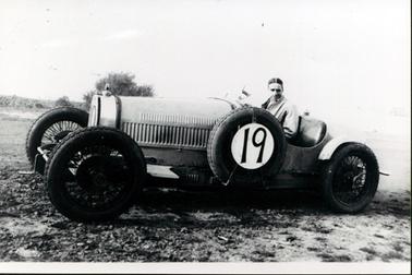 This image shows a man sitting in a open sports car smiling for the camera.  He is wearing a white shirt, tie and a light coloured coat.  His hair is parted at the centre. The spare tyre on the side of the vehicle has a large number 19 on it.  Trees can be seen in the background.   