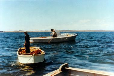 This image shows two small fishing boats and part of a third one in the foreground.  There is a man fishing in the larger boat.  He is standing underneath a shade covering.  There are white railings around the front of his boat and an anchor can be seen handing over the edge.  Another man is standing fishing in the smaller boat.  He is wearing a hat and is half turned around to look at the camera.  Fishing gear can be seen in the bottom of his boat.  Only a small section of the boat in the foreground can be seen.  Land can be seen in the background.  