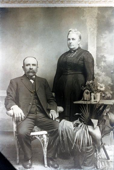 This image is a studio portrait of a man and a woman.  The man is sitting on a cane chair with his arms resting on the arms of the chair.  He is wearing a three piece suit, white shirt and bow tie.  He has a large moustache and receding hair.  The woman is standing to his right behind a small table.  A small vase of flowers is on the table.  She is wearing a long-sleeved black dress with a lace insert at the neck.  A brooch is pinned at the neckline.  Her fair hair is pulled upwards and pinned at the top.  A small potted palm tree is in the foreground on the right-hand side.  