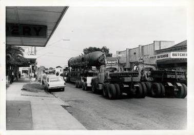 This image shows a very large cylinder being transported on a semitrailer along the street of a town.  Two trucks, with weights on the back, are pushing from the rear.  The photograph is taken from the rear of the vehicles.  Shops line either side of the street.  A line of motor cars are parked along the left-hand side of the street.  The last car is a Holden station wagon.  People can be seen standing outside the shops watching.  