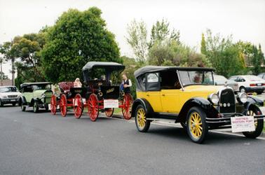 This image shows four vintage motor cars lined up on a street, waiting to proceed.  The first car is yellow with a black soft top and yellow wheel spokes.  The second car is black with a soft black top and very large red wheels.  The third vehicle is the same but without a top.  These two vehicles have the word “McIntyre” on the front in gold writing.  The last vintage car is green with a black soft top and green wheel spokes.  All cars have a sign attached to the front which reads: “Hastings-Western Port Historical Society Incorporated”.  A man can be seen sitting in the passenger seat of the yellow car and a girl in the third car.  People are standing talking beside the cars on the footpath.  Part of a car can be seen behind the green car and several cars are parked on the right-hand side of the photograph.  Trees are in the background and the bitumen street is in the foreground.  