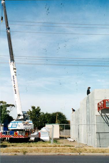 This image shows a construction site with a tall crane beside the concrete wall of a building.  Written on the crane and along the arm is: “Eazy-lift”.  “Crane Hire” is also on the side of the crane in large white letters on a blue background.  Two men can be seen on the top of the partly constructed concrete wall.  A small building is between the crane and concrete wall.  Two sets of power lines are above the building and trees are in the background.  A bitumen street is in the foreground.  