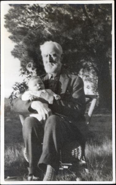 This image shows an elderly man with white hair and beard sitting on a cane chair holding a baby.  The man is dressed in a suit with his shirt buttoned up to the neck.  He has a serious expression on his face and is looking at the camera. The baby is dressed in light coloured clothes and appears to be asleep.  Untidy grass is in the foreground and a large pine tree in in the background.  