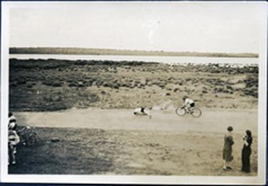 This image shows a foreshore.  Two women are standing in the foreground on the right-hand side of the image watching someone riding a bike.  Someone appears to be on the ground in front of the bike rider and a small section with children and tricycles can be seen on the far left-hand side.  Behind the bike rider are tuffs of grass, mangroves and water with a land mass in the far distance.  