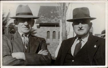 This image shows two men posing for a photograph, taken from the waist up.  They are both wearing suits, ties and hats.  The man on the right-hand side of the photograph has a darker suit and is wearing a badge on his coat lapel.  Behind them is a brick building with a steep pitched roof and three narrow windows with rounded tops.  A tree trunk is standing between the men and the building.  