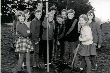 This image shows ten children and one man posing for a photograph while planting a tree.  The tree has black plastic around its base.  Everyone has big smiles.  A girl on the right, wearing a white pullover, skirt and stockings, is using a spade.  Another girl, beside the tree, is holding a long stake.  All the children are dressed in winter clothes.  Large pine trees are in the background.  