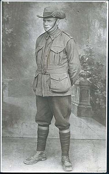 This image is of a World War 1 soldier in full army uniform posing for a studio photo.  His hands are held behind his back, his legs are parted.  