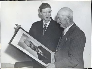 This image shows two men standing holding a framed photograph of a man, taken from the chest up.  The man on the left-hand side of the image is smiling and looking at the other man. He is wearing a dark coloured suit with vest, white shirt and tie.  He has a full head of dark coloured hair. The man on the right-hand side is looking at the photograph.  He is wearing a lighter coloured striped three-piece suit with white shirt and tie.  He has a small badge pinned to the left lapel of his coat.  He has little hair.  