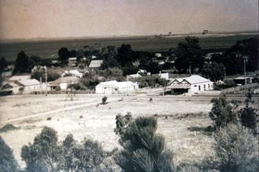 This image shows a township taken from an elevated position.  There is undeveloped land in the foreground and houses in the middle of the image, set amongst the trees.  There is a large double gabled building with a striped awning  in the centre with a street running in front of it. Trees are in the foreground and water can be seen in the background.  Land can be seen in the far background.  