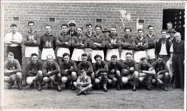 This image shows a football team posing for a photo.  There are seventeen men standing, ten men crouching and a boy sitting cross-legged at the front. The man standing on the left-hand side at the back is wearing long dark trousers, white shirt, sleeves rolled up to the elbow, and a tie.  There is a man standing behind the team, fifth from the left, wearing a cap, shirt, tie and coat and four men on the extreme right-hand side of the photograph are not wearing football uniforms.  Another man with only head and hat visible is also standing in the back row.  The are posing in front of a large brick building with four air vents visible along the wall.  