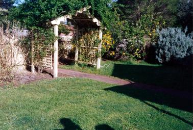 This image shows a cream coloured bower covered in a creeper.  The front of the bower has a scalloped trim and lattice is on each side.  A brick path leads to the bower.  Shrubs and trees are on either side and a green lawn is in the foreground.  Yellow fruit can be seen on a tree to the right.  A large shadow of a building can be seen on the grass on the right-hand side.  