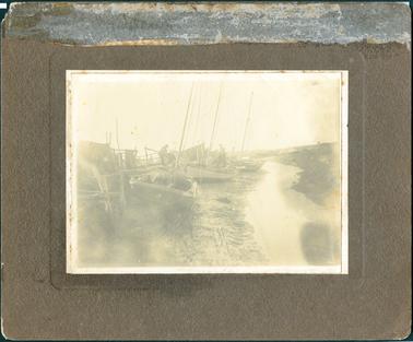 This image shows several small fishing boats on a mud bank beside a small creek.  Three of the boats have tall masts.  A man can be seen working on the middle boat and another man is nearby, slightly bent over, working on fishing gear that is hanging over a rail.  There are fences and posts on the left-hand side of the photograph with fishing nets etc hanging over them.  The two men are dressed in suits and are both wearing hats.  Small shrubs can be seen on the bank on the right-hand side of the creek.  