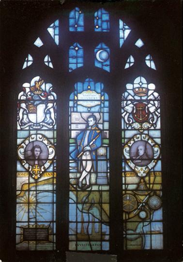This image shows a stained glass window with an image of Matthew Flinders standing in the centre panel.  He is wearing a blue and gold trimmed coat and white waistcoat and britches underneath.  He is holding a telescope in his left hand.  His right hand is on his hip and his feet are crossed.  Above him a plaque reads: Matthew Flinders 1774-1814.  On the left-hand side of the image there is an image of Joseph Banks and on the right-hand side an image of George Bass.  A map, sun dial, ship and navigating instruments are across the bottom.  Coats of arms are in the panels at the top of the window.  