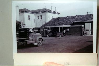 This image shows the back of a building which consists of two sections.  A tall white section with a tiled roof, two chimneys and six windows is on the left-hand side and a low, brick, tiled roof section is on the right-hand side.  A man, with his back to the camera, is walking under a small lean-to attached to the brick building.  A small door with the word ‘gents’ is on the left of the lean-to and two pot plants are at the front.  A ladder is standing against the building.  A truck, with ‘Hastings Hotel Pty Ltd’ is parked in the foreground.  