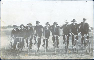This image shows a group of nine men on bicycles, in a line, posing for a photograph.  All the men are wearing suits.  Two men are wearing caps and the rest are wearing hats.  All except three are wearing ties.  They are supporting each other with hands on shoulders. They are on an open piece of ground with some buildings and trees in the far background.
