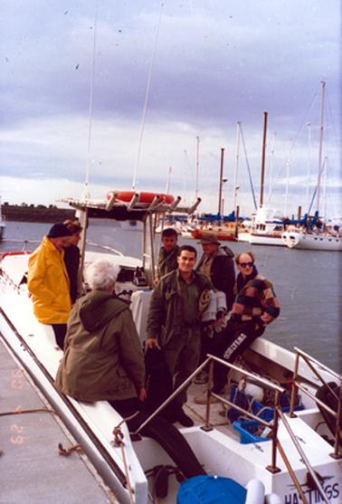 This image shows seven men standing and sitting in a boat, moored at a jetty.  Three men are on the left-hand side of the boat and four men are on the right-hand side.  The man at the centre, dressed in green is smiling for the camera.  All men are wearing jackets.  The men on the left are wearing black, yellow and green jackets and the men on the right are wearing green, black and check jackets.  The man with the check jacket has the word ‘sweathog’ on his pants in large white printing.  Equipment is on the floor of the boat in blue containers.  Five poles on a frame for holding fishing rods can be seen. The word ‘HASTINGS’ is printed on the stern of the boat. Several boats with large masts are on the right-hand side of the image.  Land can be seen in the background.  
