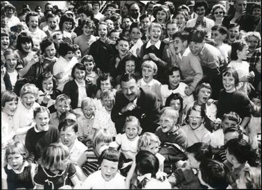 This image shows a man, dressed in a suit and tie, in the middle of a large group of over seventy children.  The children are all laughing and cheering. Adults can be seen at the back of the group.  