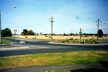 This image shows a roundabout at the right-hand side and two roads on the left-hand side.  The roundabout has a flower garden in the centre and a sign post with the number “C777” along with a smaller white street sign.  Tall power poles and electricity wires and a street lamp are on each corner.  The paddock in the background has several large bales of hay lying on the ground.  Trees are scattered across the paddock and the roofs of houses are in the far background.  A grassed and paved footpath are in the foreground.  