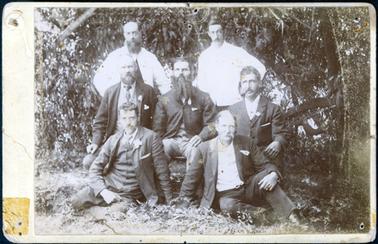 This image shows a group of seven men posing for a photograph.  Two men are standing at the back, wearing white coats.  Three men are sitting in the centre row and two men are sitting on the grass in the front.  The men in the centre and front rows are wearing suits. All men have moustaches and three have beards as well. The man in the centre of the photograph has a very long beard.  None of the men are smiling.  Low hanging branches are behind the group.  