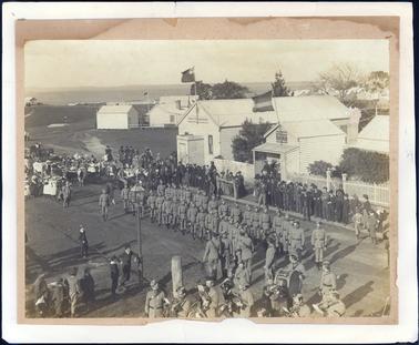 This image is of a military detachment marching in the street led by a military band.  A soldier on horseback is behind them and a team of bullocks and more horses are following.  Men, women and children are lining the street watching the parade.  Everyone is very well dressed.  Some of the children are waving flags.  A cast iron gas lamp is in the foreground.  Wooden shops and buildings with iron roofs line the street, including the Library and Post Office, both of which are flying flags.  A white picket fence is seen on the right-hand side of the image.  Many other buildings are seen in the distance and the Western Port, looking to French Island is seen in the background.  A fishing shed can be seen at the top left-hand side.  A hitching rail is in front of the Post Office.  