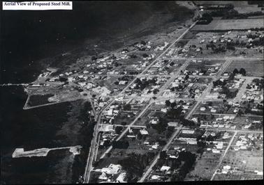 This image shows an aerial photograph of a town with the foreshore and jetties on the left-hand side.  Many houses, streets and trees can be seen.  Some rural paddocks can be seen at the top and on the far right-hand side of the image.  A piece of white paper with the words “Aerial View of Proposed Steel Mill.” typed in black, is glued to the top left-hand corner of the image.  