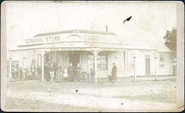 This image shows the general store in Hastings. It is a timber building with a brick roof and a verandah with a corrugated iron roof. The verandah posts are decorated at the top. At the top of the verandah is a large sign with the words “GENERAL STORE” written twice and the words “H. PROSSER” in between. Below the verandah roof around its extent are the words “flour bran & pollard oats maize & chaff drapery boots & shoes groceries”. The residential dwelling is attached to the right side of the building. Under the verandah to the left is a small group of children. Under the verandah in front of the building are 4 adults and 5 children, and to their right is a man and a dog. The floor of the verandah and the ground around is dirt. On the back of the image is written in biro “Hastings General Store Mr & Mrs Harry Prosser”