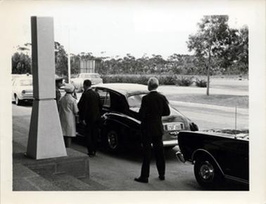 This image shows two men and a woman standing beside a car with their backs to the camera.  Part of a man in uniform can be seen beside the woman.  The two men are dressed in dark coloured suits.  The woman is wearing a coat and hat.  The car has the registration number of ZSF-200.  The car is parked in front of a building. Part of a memorial is seen in the foreground on the left-hand side of the image.  Four other cars can be seen.  There are gardens and trees in the background.  