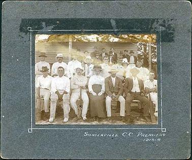 This image shows some members of a cricket team along with a woman and other men.  They are sitting and standing in front of a wooden pavilion.  Members of the public are seen standing at the back of the group.  A large pine tree is on the right-hand side at the back. The inscription on the front is “Somerville C.C. Premiers 1913-14.” 