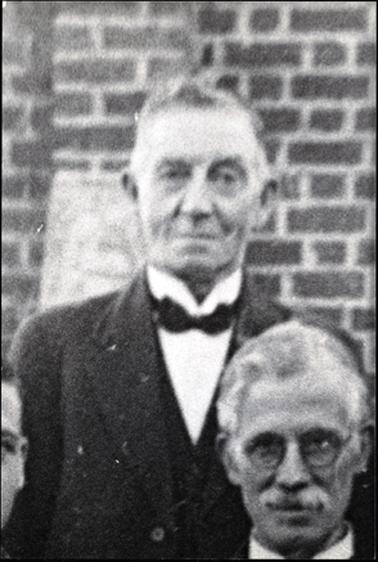 This image shows the top half of a man standing looking at the camera.  He has a slight smile on his face. He is wearing a three piece suit, a white shirt with a stand-up collar and a black bow tie.  A brick wall is behind him.  The head of a man with white hair, a moustache and spectacles is on the right-hand side of the image and part of a head can be seen on the left-hand side of the image.  