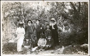 This image shows a group of nine people posing for a photograph in bushland.  Two woman and three men  are standing and the back with a small boy standing in front of them.  Two men and a woman are sitting in the front on the grass.  The woman on the left-hand side of the image is wearing a white long sleeved outfit with an overlay on each side of the skirt.  Her hat has a dark band with flowers.  The woman on the right-hand side is wearing a black suit with a white blouse.  Her hat has protective netting coming around her shoulders.  The woman sitting on the grass has a light coloured coat over her clothes and a large dark hat.  All the men are wearing three-piece suits, hats and have beards and moustaches.  The boy has short, fair hair and is dressed in a sailor suit.  There is bushland all around them and bracken fern can be seen in the foreground.  Part of a horse can be seen in the background.  