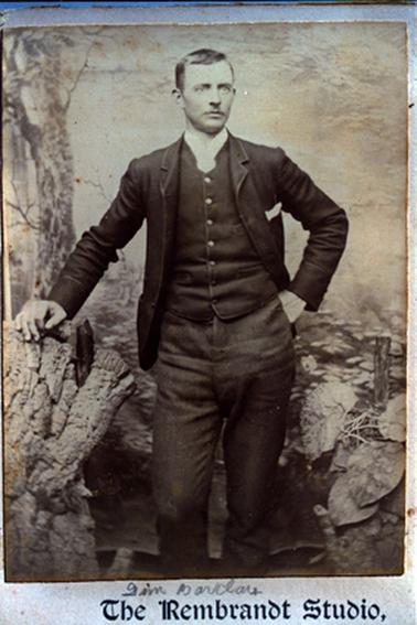 This image shows a young man standing beside a tree trunk, posing for a photo.  He is looking slightly away from the camera.  He is wearing a buttoned-up waistcoat, jacket, lighter coloured trousers and a high necked white shirt.  A white handkerchief is in the left side pocket of his jacket.  His right hand is resting on the stump and his left hand is in his trouser pocket.   Printed across the bottom of the photograph is: “The Rembrandt Studio.”.  