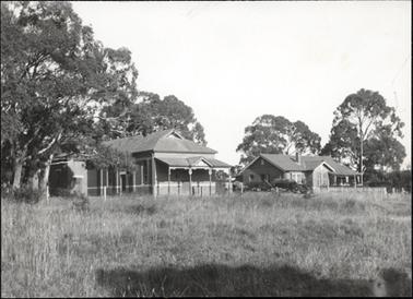 This image is of the Catholic School in Hastings.  There are two brick buildings and both have triangular pediments at the front along with verandah posts.  The building on the left of the photograph has a white line running around it about one third up from the ground.  Both buildings appear to have tiled roofs.  Two brick chimneys are seen on the building on the right of the photograph.  There is a large expanse of grass in the foreground and large trees are seen on both sides of the buildings and at the back of the buildings. 