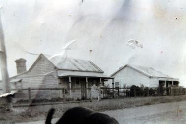 This image shows two weatherboard  cottages with sloping roofs.  The cottage on the left has a four posted verandah across the front.  There are two windows on either side of the centre front door.  One window is on the side of the building and part of a large chimney can be seen at the back.  The building on the right has a three posted verandah across the front and a small window on the side.  A front fence stretches across the front of the buildings and a grassy footpath and a gravel road is in the foreground.  Part of a dog can be seen at the bottom of the image.  