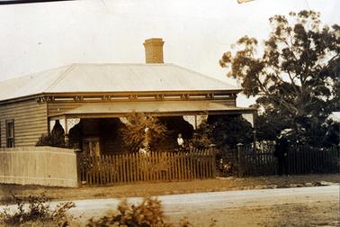 This image shows a pretty weatherboard house with a sloping roof and a four post ornamental verandah across the front.  A lace curtain can be seen behind the window on the front left-hand side of the house.  Two windows can be seen along the side.  A large chimney can be seen at the rear of the house.  A wooden fence runs along the side of the house and a wooden picket fence runs across the front.  A small gate at the centre is open.  A well established garden can be seen in the front yard behind the fence.  A man is standing on the footpath at the right of the gate and a woman is standing on the verandah.  A tall tree on at the far right-hand side of the house.  A grassed footpath and a gravel road is in the foreground.  