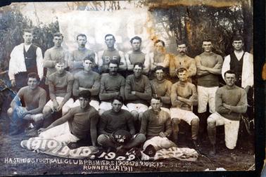 This image shows a football team posing for a photo.  There are nine men standing in the back row, eight men sitting in the middle row and three men sitting on the ground in the front row.  The players are wearing either high-necked longsleeve or sleeveless t-shirts.  Their knee length shorts are white.  The two men on either end of the back row are wearing long sleeved white shirts with a dark coloured vest and have a towel over their shoulder.  The centre player in the front row is holding a ball with ‘H.F.C.’ printed in white.  A large banner with the letters ‘H.F.C.’ is behind the group and a long narrow banner with numbers printed across it is on the ground in front of the group.