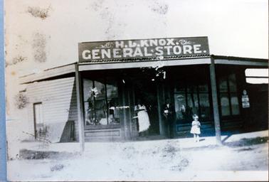 This image show a  weatherboard building with a large signed on the verandah roof which reads, ‘H.L. Knox General Store’. The verandah has two posts on the footpath and a door in the centre with large windows on either side.  A small door is along the left-hand side of the building.  Two people are standing at the front door and a small girl is standing on the footpath.