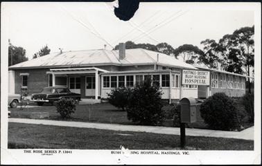 This image shows the front and side view of a building with the sign ‘Hastings District Bush Nursing Hospital’ in the grounds on the right-hand side of the image.  The building is brick construction with an iron roof.  A two post skillion is over the large double doors at the front of the building.  Sets of windows are on the right-hand side at the front and down the side.  A dark coloured motor car and part of another car are parked in front of the building.  There is a large lawn with shrubs at the front and side, a concrete footlpath and a lawned footpath.  A mail box is on the footpath in the foreground on the right-hand side.  The tops of large trees are behind the building.  The following is printed across the bottom: “The Rose Series P. 13941 (copyright) Bush Nursing Hospital, Hastings, Vic.”.  A round piece is torn away at the centre top of the postcard and there are scratches across the top.
