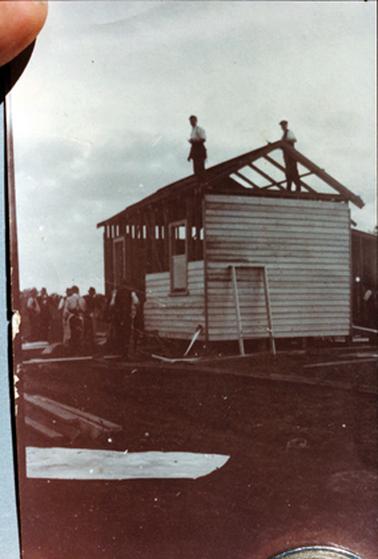 This image shows a partly built house with two men standing on the roof trusses and a group of approximately seven men standing beside the house.  No iron is on the roof but the end wall is clad in weatherboard as well as part of the side.  Two window frames are along the side wall and another frame is standing against the side wall.  Building materials are spread around the ground.  