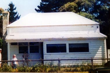 This image shows a timber house with a galvanized iron roof behind a wire mesh fence.  A large brick chimney is on the far left-hand side of the building.  A small porch with a set of square windows and one verandah post is on the left.  Two wide windows are on the right-hand section of the house.  Tall trees are behind the house and small shrubs are along the fence line.  A man and child are standing in the front garden looking at the camera.  