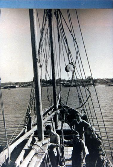 This image shows the stern of a wooden sailing vessel with four men on board. The men are all looking to the left hand side of the boat. The man at the centre of the boat is wearing a hat. The sails are down. Buildings can be seen in the background and trees in the far background.
