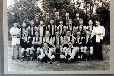 Image depicts a football team standing and sitting in four rows posing for a photograph. There are five men in  the back row, ten men standing in the second back row, seven men sitting on chairs in the front row and two men sitting crossed legged on the grass. The men in the back row are all wearing three piece suits. The man sitting at the centre in the front row, is holding a football. Tall trees are behind the men.