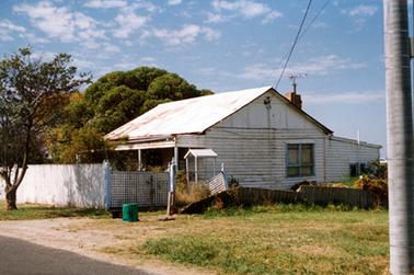 This image shows a side-on view of a timber home with a galvanized iron roof.  A four post verandah stretches across the front.  A large window is along the side of the house and a lean-to is at the back.  Part of a brick chimney with a television antennae can be seen on the roof.  A tall paling fence is at the front of the house and a panel of lattice fencing is along the side as well as a timber fence which has partly fallen over.  A tree is on the footpath and thick trees are on the left-hand side of the house.  Part of a bitumen road and untidy grass is in the foreground.  