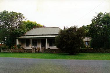 This image shows a timber house with a galvanized iron roof and a four post verandah across the front.    A large set of windows are on either side of the front door.  A low brick fence runs across the front between the house and the green grassed footpath.  A bitumen road is in the foreground.  A tree is on the footpath and large trees are on either side of the house.  Part of a building showing a window can be seen between the trees on the right-hand side of the image.  Colourful shrubs are behind the fence on the far left-hand side.  