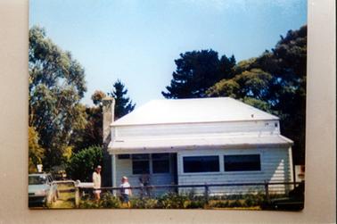 This image shows a timber house with a galvanized iron roof behind a wire mesh fence.  A large brick chimney is on the far left-hand side of the building.  A small porch with a set of square windows and one verandah post is on the left.  Two wide windows are on the right-hand section of the house.  Tall trees are behind the house and small shrubs are along the fence line.    A man and child are standing in the front garden looking at the camera.  Part of a blue motor car can be seen behind a small gate which has a letter box on the post.