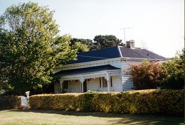 This image shows a white weatherboard house with a grey sloping roof and verandah held up by four ornate posts and wrought-iron lace.  Windows are on either side of the central front door.  A brick chimney is on the right-hand side of the roof and a television antenna is beside it.  A neatly trimmed hedge forms the front fence and a letter box is on the post beside a small white paling gate.  Tall trees are on the left-hand side and behind the house.  Smaller trees are on the right-hand side.  A grassed footpath is in the foreground. 