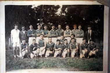 This image shows a football team posing for a photo.  There are eleven men standing in the back row and eleven men kneeling in the front row.  All the men except three in the back row, are dressed in a football uniform of white shorts and green shirts.  One of the players in the front row is holding a ball. None of the men are smiling.  Tall trees are behind the group and grass is in the foreground. 