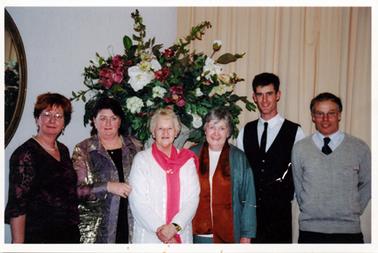 This image shows a group of four women and two men, all well dressed, standing in a line, posing for a photo.  There are four women on the left and two men on the right.  A very large flower arrangement is behind the group and a full-length cream curtain is on the wall on the right-hand side.  
